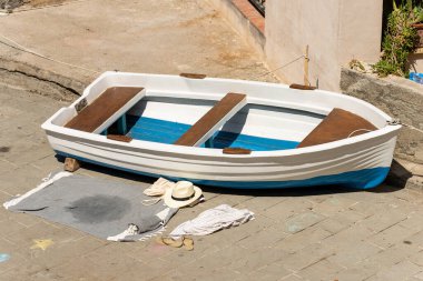 Small white and blue rowing boat moored on the quay of the port, a beach towel, sun hat and flip-flop. Tellaro village, tourist resort in La Spezia province, Liguria, Italy, southern Europe.