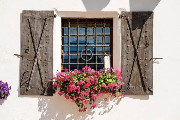 Close-up of an ancient window with wrought iron security bars, metal shutters and red geranium flowers. Small village of Malborghetto-Valbruna, Udine province, Friuli-Venezia Giulia, Italy, Europe.