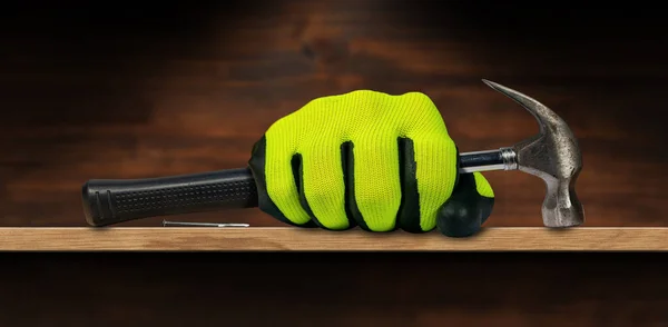 Hand with green and black protective work glove holding an old steel claw hammer with black rubber handle, on a wooden workbench with a nail and copy space.