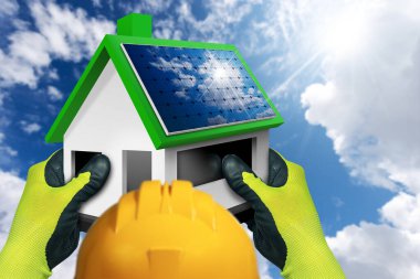 Koruyucu iş eldivenleri ve turuncu şapkalı inşaat işçisi çatıda güneş panelleri olan küçük bir model ev tutuyor, arkasında bulutlar ve güneş ışınları olan mavi bir gökyüzü. Yenilenebilir enerji kavramı