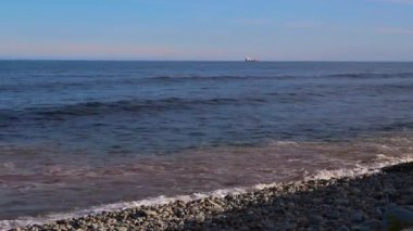 Kıyıda köpük ve sprey ile deniz dalgası ve ufukta kargo gemisi.