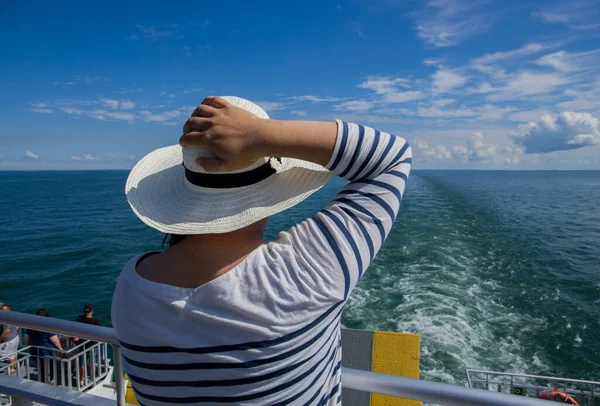 A woman in a hat stands at the stern of a ship and looks at the sea. Sunny day, cloudy sky, blue tones