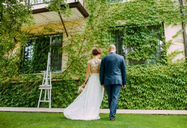 Mutlu yeni evliler el ele tutuşup çimlerde yürür ve lianaların eviyle iç içe geçerler. Arka plan.