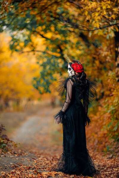 Young woman with sugar skull makeup and red roses dressed in black costume of death as Santa Muerte is against background of autumn forest. Day of the Dead or Halloween concept.