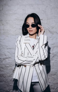 Güneş gözlüklü şık genç kadın, çizgili beyaz ceket ve şort giymiş tuğla duvarın yanında el çantasıyla duruyor..