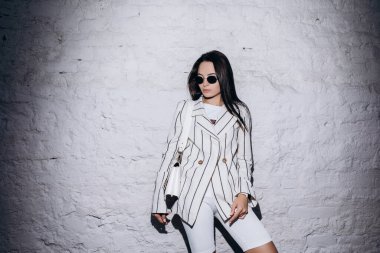 Güneş gözlüklü şık genç kadın, çizgili beyaz ceket ve şort giymiş tuğla duvarın yanında el çantasıyla duruyor..