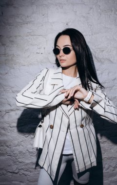 Güneş gözlüklü şık genç kadın, çizgili beyaz ceket ve şort giymiş tuğla duvarın yanında el çantasıyla duruyor..