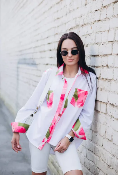 Güneş gözlüklü şık genç bir kadın renkli şeffaf bir bluz ve beyaz şortla tuğla duvar arkasında duruyor..