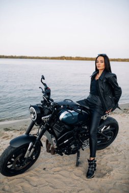 Genç bir kadın nehir kıyısındaki kumlu sahilde deri ceketli ve pantolonlu siyah bir motosikletin üzerinde oturuyor..