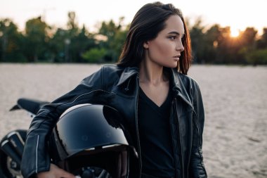 Deri ceketli genç bir kadın elinde miğferiyle kumlu plajın ortasında gün batımında siyah motosikletin arka planına karşı duruyor. Portre.