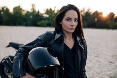 Deri ceketli genç bir kadın elinde miğferiyle kumlu plajın ortasında gün batımında siyah motosikletin arka planına karşı duruyor. Portre.