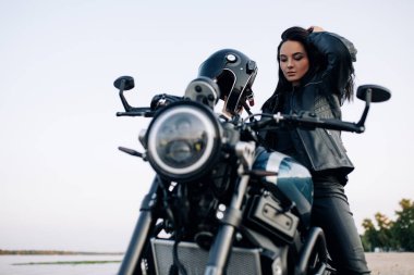 Genç bir kadın nehir kıyısındaki kumlu sahilde kasklı ve deri ceketli siyah bir motosiklette oturuyor..