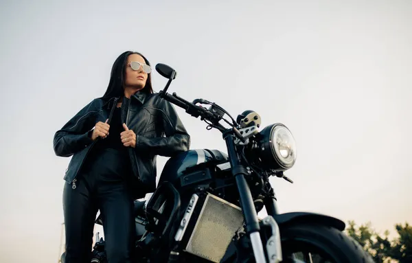 年轻的黑发女子站在黑色摩托车旁 戴着太阳镜 皮夹克和裤子 背对着天空 — 图库照片