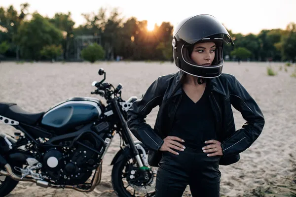 若いです女性はヘルメットと革のジャケットの中で白いバイクの背景を背景に砂浜日没で立ちます — ストック写真