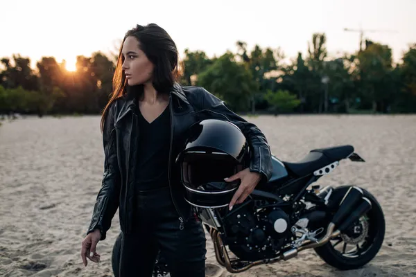 若いです女性スタンドで革のジャケットでヘルメットで彼女の手の中に砂浜の間で黒バイクの背景に夕日 — ストック写真