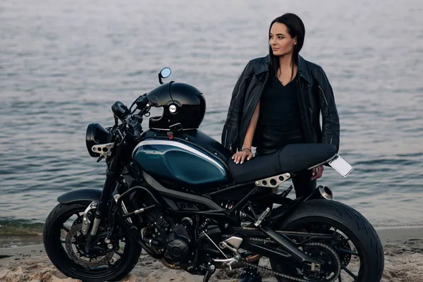 若い女性は川の海岸近くの砂浜で革のジャケットとパンツの黒いバイクの横に立つ — ストック写真