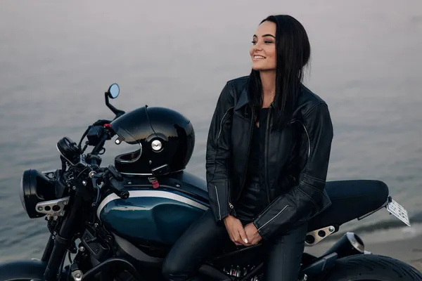 年轻女子站在身穿皮夹克和裤子的黑色摩托车旁边 靠着河边 — 图库照片