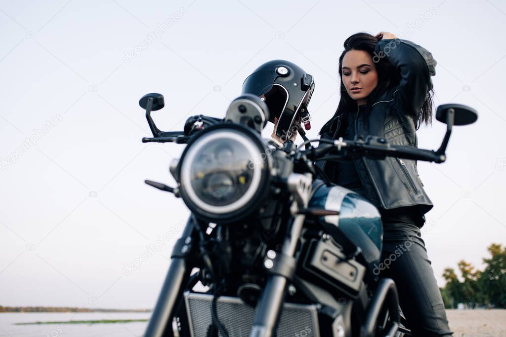 Young woman sits on black motorbike with helmet and leather jacket on sandy beach next coast of river.