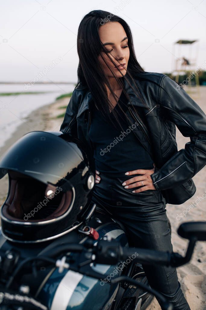 Young woman sits on black motorbike in leather jacket and pants on sandy beach next coast of river. Closeup.