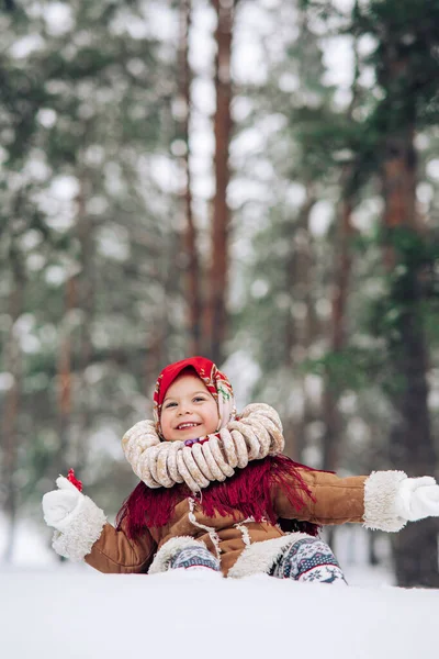 Beautiful child girl has fun with lollipop in hand and bagels bunch on her neck in snowy forest. She dressed in the old Russian style in red headscarf.