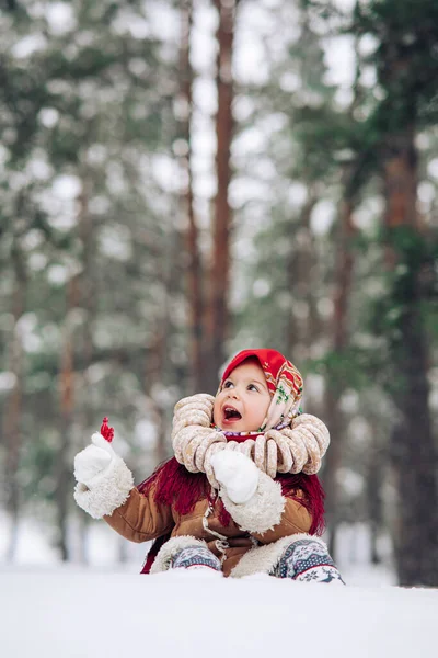 Beautiful child girl has fun with lollipop in hand and bagels bunch on her neck in snowy forest. She dressed in the old Russian style in red headscarf.