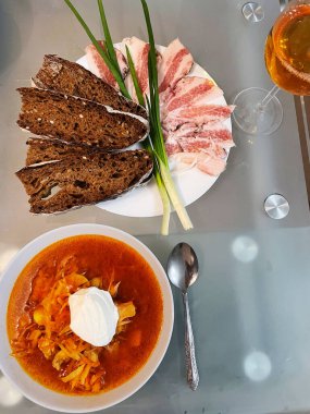 Ekşi kremalı borş soslu bir tabağın üst görüntüsü, çavdar ekmeği dilimleri, domuz yağı ve masada duran yeşil soğan tüyleri..