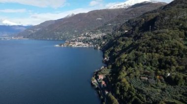 İtalya Gölü 'nün su yüzeyinden güzel drone manzarası Sahil boyunca Bellano' dan Bellano 'ya, mavi gökyüzü, bulutlar ve karla kaplı Alpler uzaktaki tepeler.
