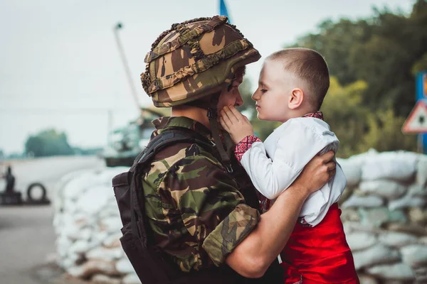 Ukrainian soldier holds child boy in traditional national dress in his hands on roadblock against background of sandbags. Concept of russian military invasion in Ukraine. War in Ukraine and Europe. Children and war.