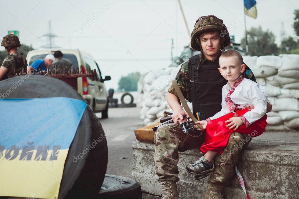 Ukrainian child boy in traditional national dress sits near ukrainian soldier on roadblock against background of sandbags. Concept of russian military invasion in Ukraine. War in Ukraine and Europe. Children and war.