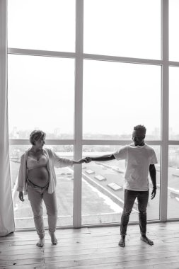 Genç, mutlu Afrikalı bir adam ve beyaz hamile bir kadın pencerenin önünde el ele tutuşuyorlar. Irklar arası evlilik kavramı. Siyah ve beyaz resim.