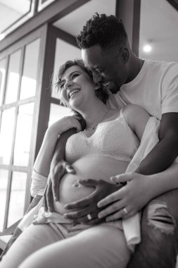 Genç, mutlu Afrikalı bir adam ve beyaz hamile bir kadın birlikte oturur, eğlenir ve neşeyle gülerler. Irklar arası evlilik kavramı. Siyah ve beyaz resim.