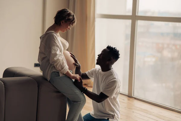 若い幸せなアフリカ人男性は 白人の妊婦の前でひざまずき 彼の手で彼女の腹に触れます 異人種間結婚の概念 — ストック写真
