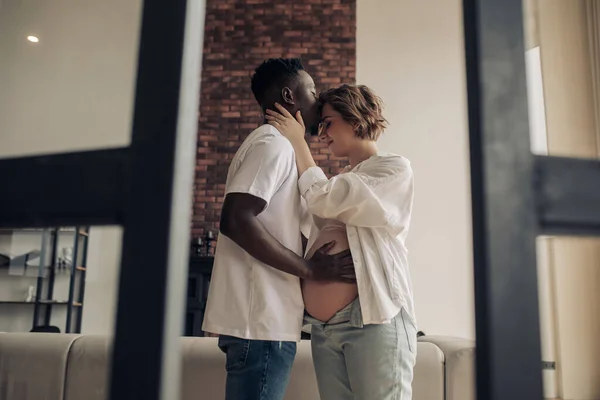 若い幸せなアフリカ人男性が立って 包含し 彼の白人妊娠中の女性にキスし 彼の手で彼女の腹に触れます 異人種間結婚の概念 — ストック写真