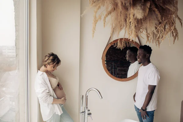 若い幸せな白人妊娠中の女性が立っており 窓の背景に対して浴室でアフリカ人男性の横に彼女の腹に触れます 異人種間結婚の概念 — ストック写真
