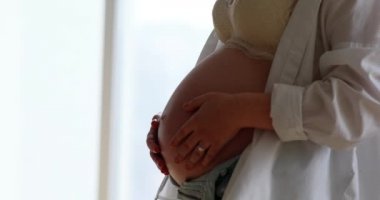 Hamile kadın odanın penceresinin yanında duruyor, elleriyle karnına dokunup okşuyor. Annelik ve hamilelik kavramı. Yakın çekim..