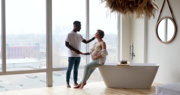 年轻快乐的非洲男人手牵手 坐在浴室边缘 靠窗的背景的高加索孕妇 种族间婚姻的概念 产妇和怀孕 — 图库视频影像
