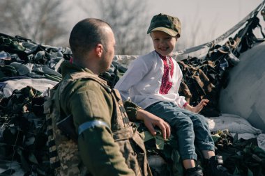 Nakışlı tişörtlü çocuk Ukrayna bölge savunma savaşçısı yakınlarındaki kontrol noktasında kum torbalarının üzerinde oturuyor. Ukrayna 'da Rus askeri istilası kavramı. Ukrayna ve Avrupa 'da savaş. Çocuklar ve savaş.