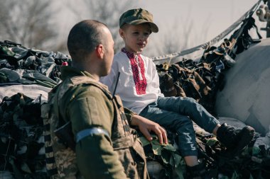 Nakışlı tişörtlü çocuk Ukrayna bölge savunma savaşçısı yakınlarındaki kontrol noktasında kum torbalarının üzerinde oturuyor. Ukrayna 'da Rus askeri istilası kavramı. Ukrayna ve Avrupa 'da savaş. Çocuklar ve savaş.