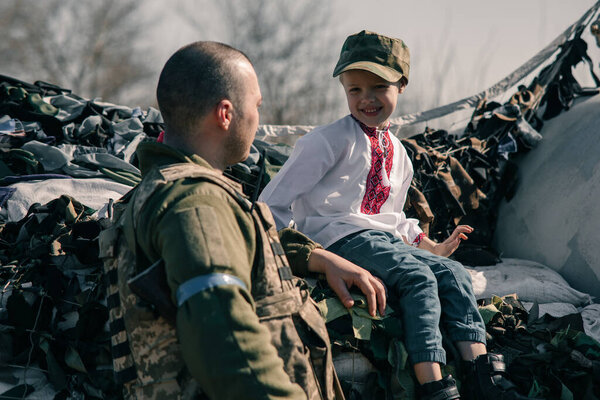 Child boy in embroidered shirt sits on sandbags in checkpoint near Ukrainian territorial defense warrior. Concept of russian military invasion in Ukraine. War in Ukraine and Europe. Children and war.