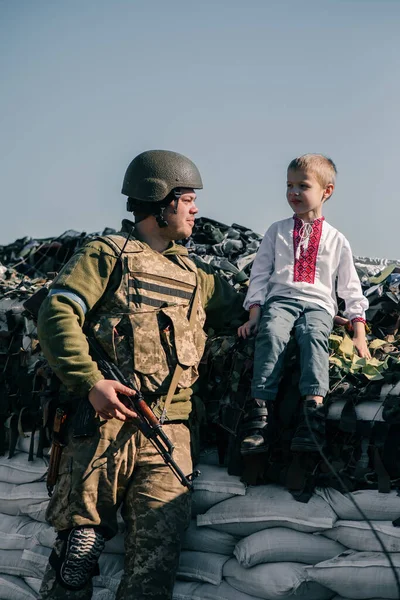 Child boy in embroidered shirt sits on sandbags in checkpoint near Ukrainian territorial defense warrior. Concept of russian military invasion in Ukraine. War in Ukraine and Europe. Children and war.