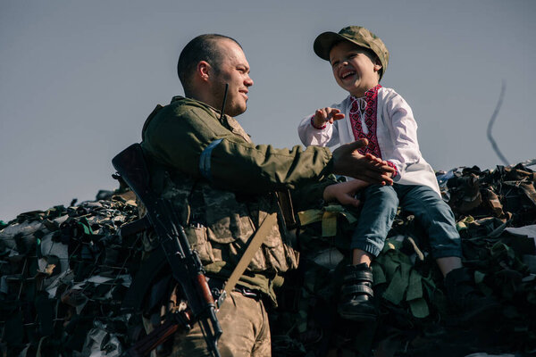 Child boy in embroidered shirt sits on sandbags and laughs in checkpoint near Ukrainian territorial defense warrior. Concept of russian military invasion in Ukraine. War in Ukraine. Children and war.