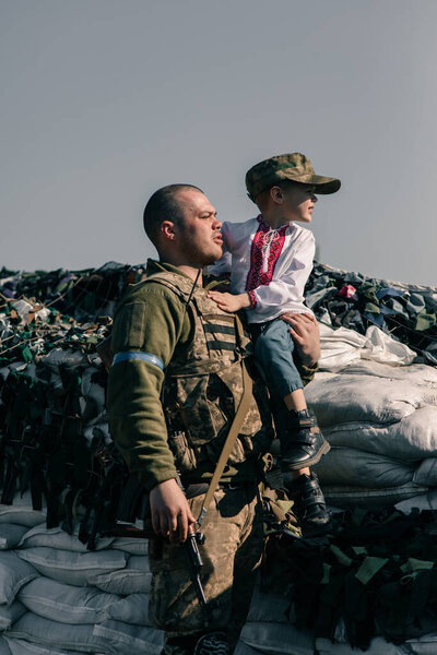 Ukrainian territorial defense warrior holds child boy in embroidered shirt in his hands on checkpoint. Concept of russian military invasion in Ukraine. War in Ukraine and Europe. Children and war.