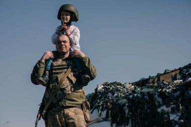 Omzunda çocuk olan Ukraynalı asker kum torbalarıyla kontrol noktası yakınında duruyor. Ukrayna 'da Rus askeri istilası kavramı. Ukrayna 'da savaş. Çocuklar ve savaş. Boşluğu kopyala.