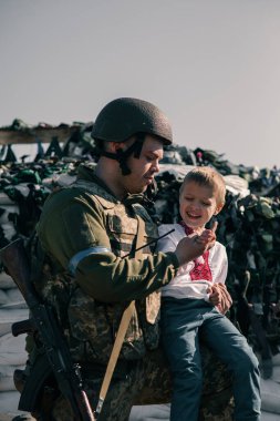Nakışlı gömlekli çocuk, kum torbalarına karşı kontrol noktasında Ukrayna bölge savunma savaşçısının yanında oturuyor. Ukrayna 'da Rus askeri istilası kavramı. Ukrayna ve Avrupa 'da savaş. Çocuklar ve savaş.