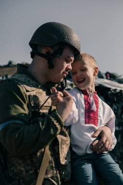 Ukraynalı asker kontrol noktasında yanında oturan nakışlı gömlekli çocuğu kucaklıyor. Ukrayna 'da Rus askeri istilası kavramı. Ukrayna ve Avrupa 'da savaş. Çocuklar ve savaş.