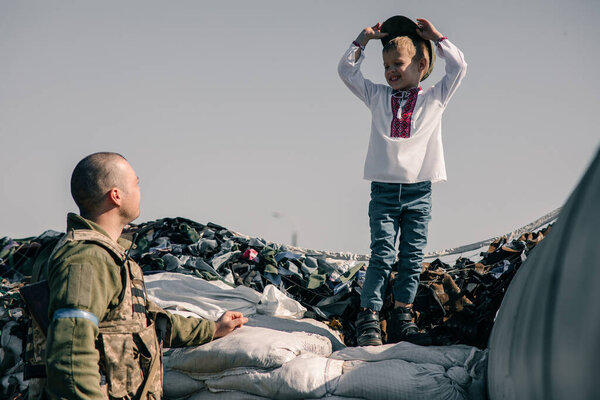 Child boy in embroidered shirt stands on sandbags in checkpoint near Ukrainian territorial defense warrior. Concept of russian military invasion in Ukraine. War in Ukraine and Europe. Children and war