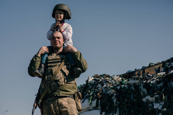 Ukrainian soldier with child boy on his shoulders stands near checkpoint against sandbags background. Concept of russian military invasion in Ukraine. War in Ukraine. Children and war. Copy space.