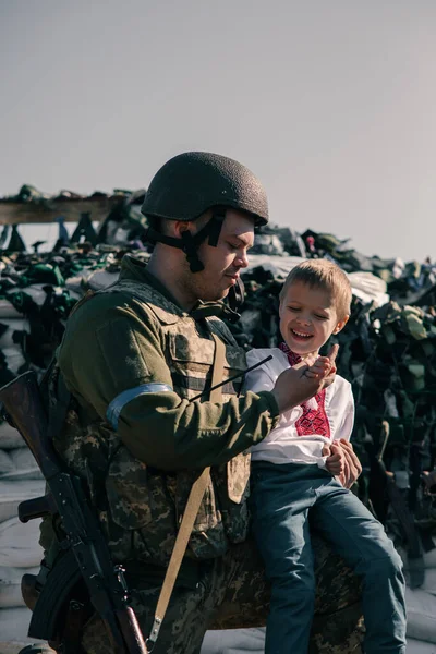 Child boy in embroidered shirt sits near Ukrainian territorial defense warrior on checkpoint against sandbags background. Concept of russian military invasion in Ukraine. War in Ukraine and Europe. Children and war.
