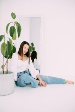 Genç esmer kadın çiçek saksısının yanında oturuyor. Çiçek saksısı aynadaki yansımasının arka planında. Beyaz arkaplan izole görüntü.
