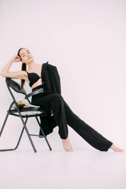 Siyah ceketli, pantolonlu ve sütyenli genç esmer kadın sandalyede çıplak ayakla oturuyor. Beyaz arkaplan izole görüntü.
